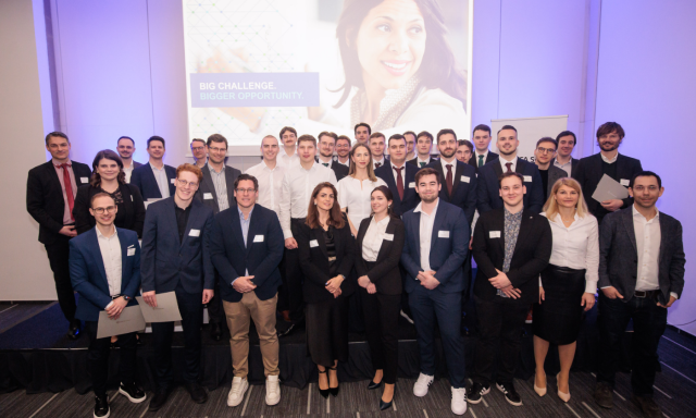 Velika skupina profesionalno oblečenih ljudi pozira skupaj za fotografijo na odru, za njimi pa je zaslon z napisom »Veliki izziv. Še večja priložnost«.
