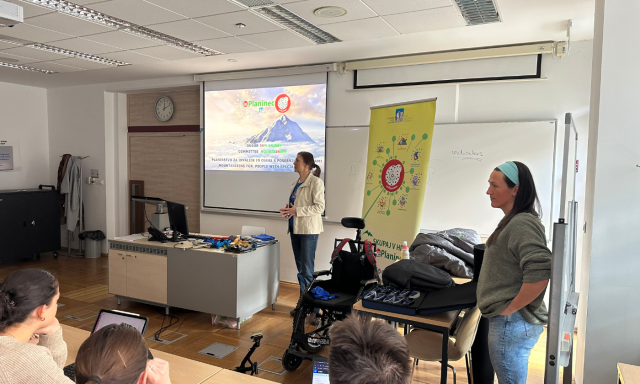A woman presents in a classroom with a projector displaying "Planinec." A banner stands beside her. Several students focus on the presentation.