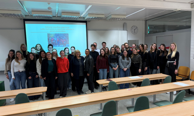 Skupina študentov in profesorjev pozira v učilnici. Stojijo pred zaslonom, na katerem je prikazana barvita diapozitiva, ki izraža občutek dosežka.