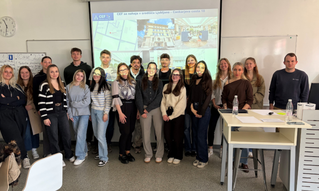 V učilnici stoji skupina ljudi in se nasmiha. Za njimi so na projekcijskem zaslonu prikazane slike stavbe in zemljevida. Vzdušje je prijazno in sodelovalno.