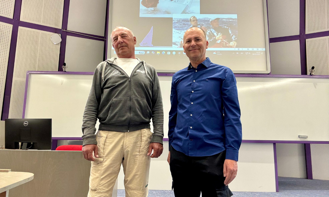 Two men stand in a lecture room, one in a grey hoodie, the other in a blue shirt. Behind them, a screen displays sailing images, suggesting a presentation setting.