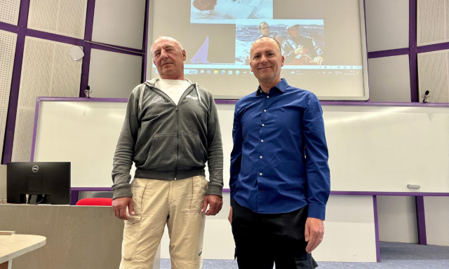 Two men stand in a lecture room, one in a grey hoodie, the other in a blue shirt. Behind them, a screen displays sailing images, suggesting a presentation setting.