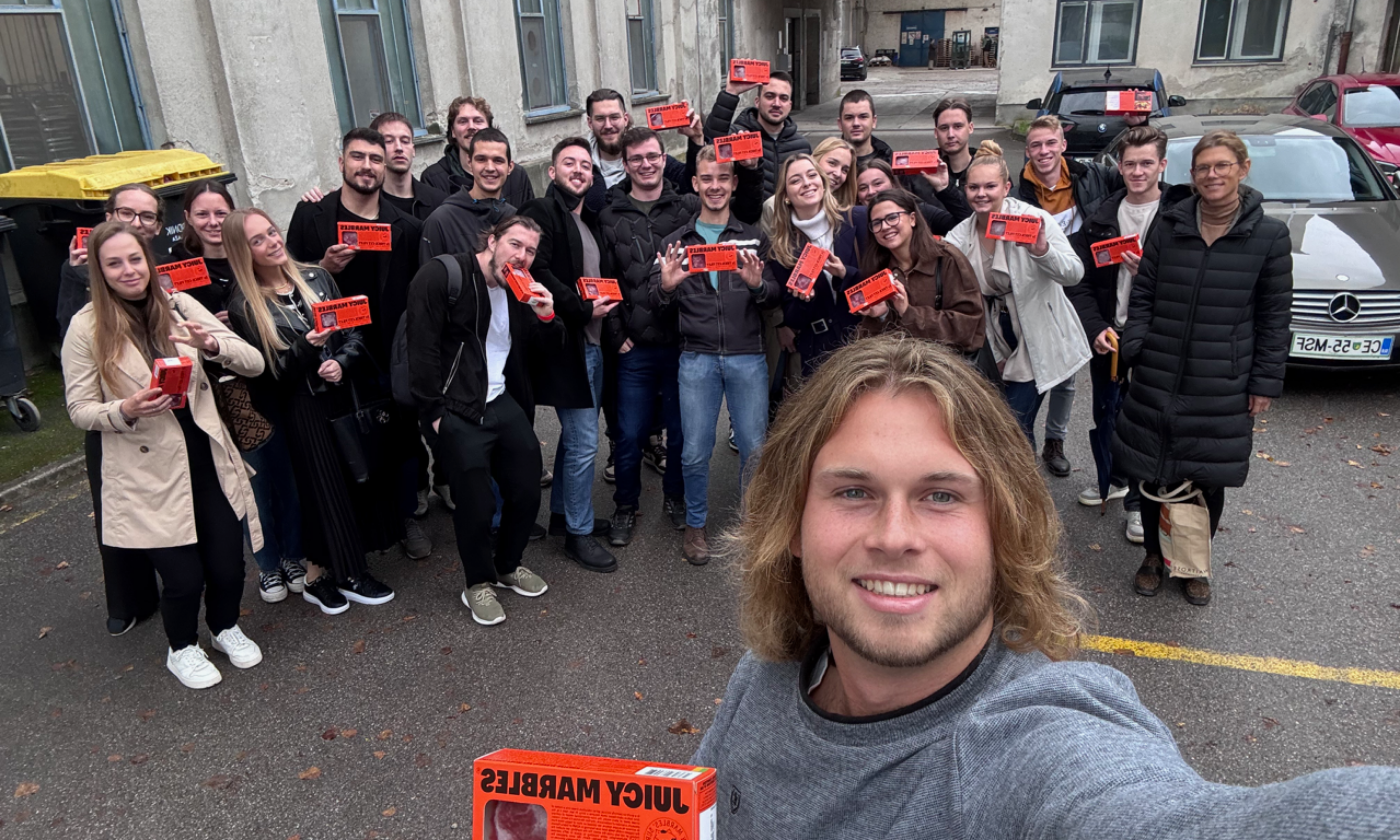 Skupina nasmejanih ljudi pozira za selfi na prostem, vsak z jasno oranžno škatlo »Juicy Marbles« v rokah. V ozadju je vidna stavba in parkirana vozila, kar ustvarja veselo in praznično vzdušje.