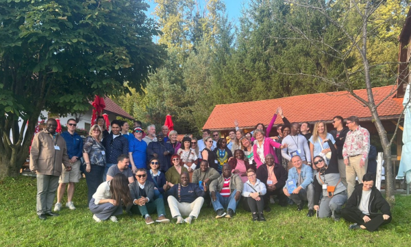Velika skupina odraslih pozira za fotografijo na prostem na travi, v ozadju pa so drevesa in stavbe z rdečimi strehami na sončen dan.