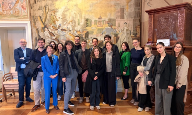 A group of 15 people stands together in a room with a large, detailed mural on the wall. The group appears cheerful and well-dressed, suggesting a formal occasion or event.