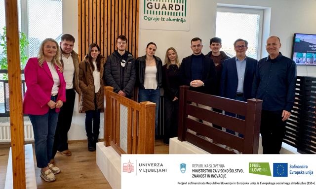 Raznolika skupina ljudi stoji skupaj pred lesenim znakom, se smeji in pogovarja med seboj.