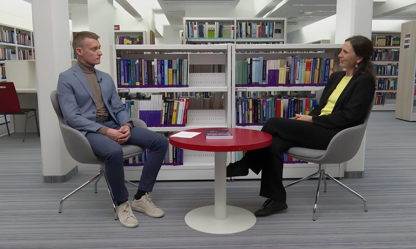 Dve osebi sedita v knjižnici in se pogovarjata za rdečo mizo. Knjižne police, polne barvitih knjig, tvorijo ozadje in ustvarjajo učenjaško vzdušje.