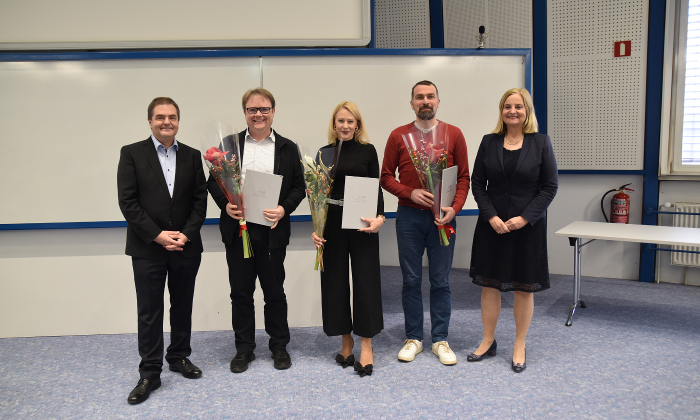 Pet ljudi stoji v vrsti in se smeji. Trije v sredini držijo certifikate in cvetje. Prizor izraža občutek praznovanja in dosežka.