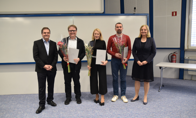 Pet ljudi stoji v vrsti in se smeji. Trije v sredini držijo certifikate in cvetje. Prizor izraža občutek praznovanja in dosežka.