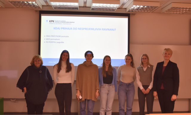 Skupina sedmih ljudi stoji pred projekcijskim zaslonom v učilnici. Na zaslonu je prikazan tekst v tujem jeziku. Skupina deluje profesionalno in mirno.