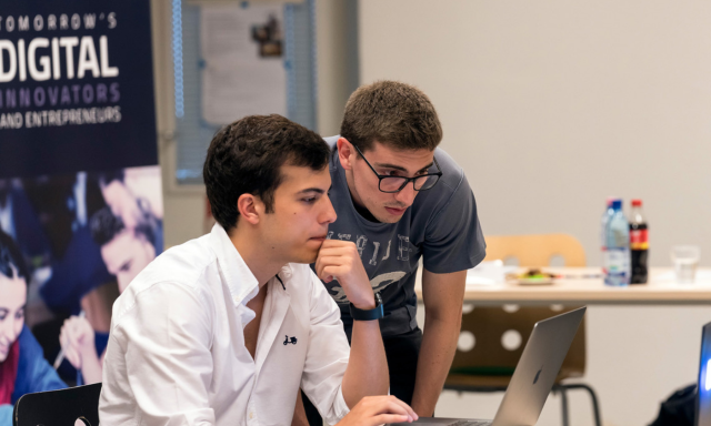 Dva mlada moška sta osredotočena na zaslon prenosnega računalnika v sodobnem delovnem prostoru, v ozadju pa sta pijača in promocijski plakat.