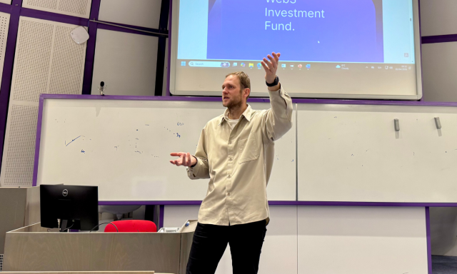 A man in a beige shirt is giving a presentation in a classroom with a projector displaying smoothie images. He appears engaged and confident.