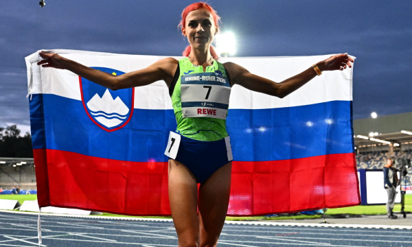 Atletinja Klara Lukan stoji na stezi in ponosno drži slovensko zastavo nad glavo.