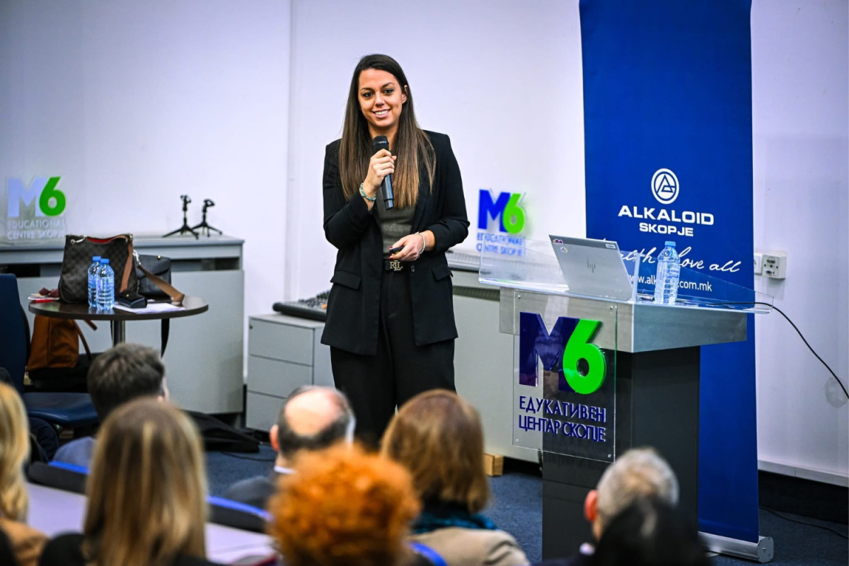 A woman in a black suit speaks into a microphone at a podium during a presentation in a conference room, with an audience seated in front of her.