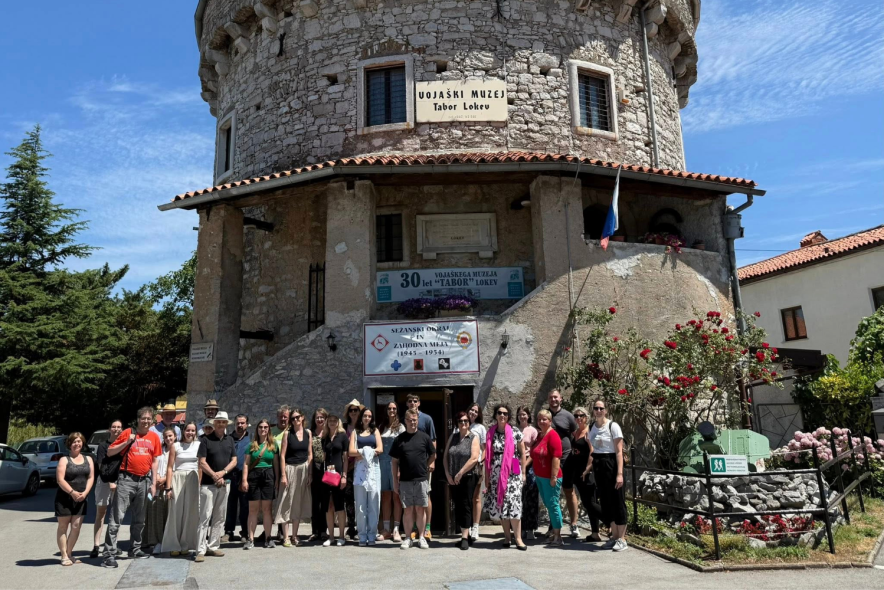 Skupinska fotografija ljudi pred kamnitim stolpom v kraški vasici. 