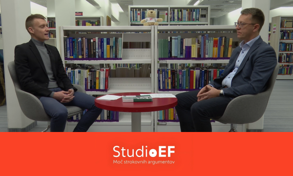 Dva moška sedita na stolih, drug nasproti drugega, v ozadju pa so knjižne police, polne knjig.