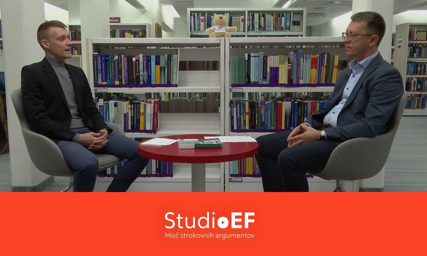 Dva moška sedita na stolih, drug nasproti drugega, v ozadju pa so knjižne police, polne knjig.