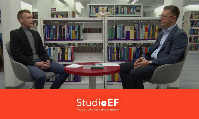 Dva moška sedita na stolih, drug nasproti drugega, v ozadju pa so knjižne police, polne knjig.