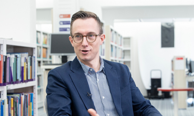 A man wearing glasses sits in front of a large bookshelf filled with various books.