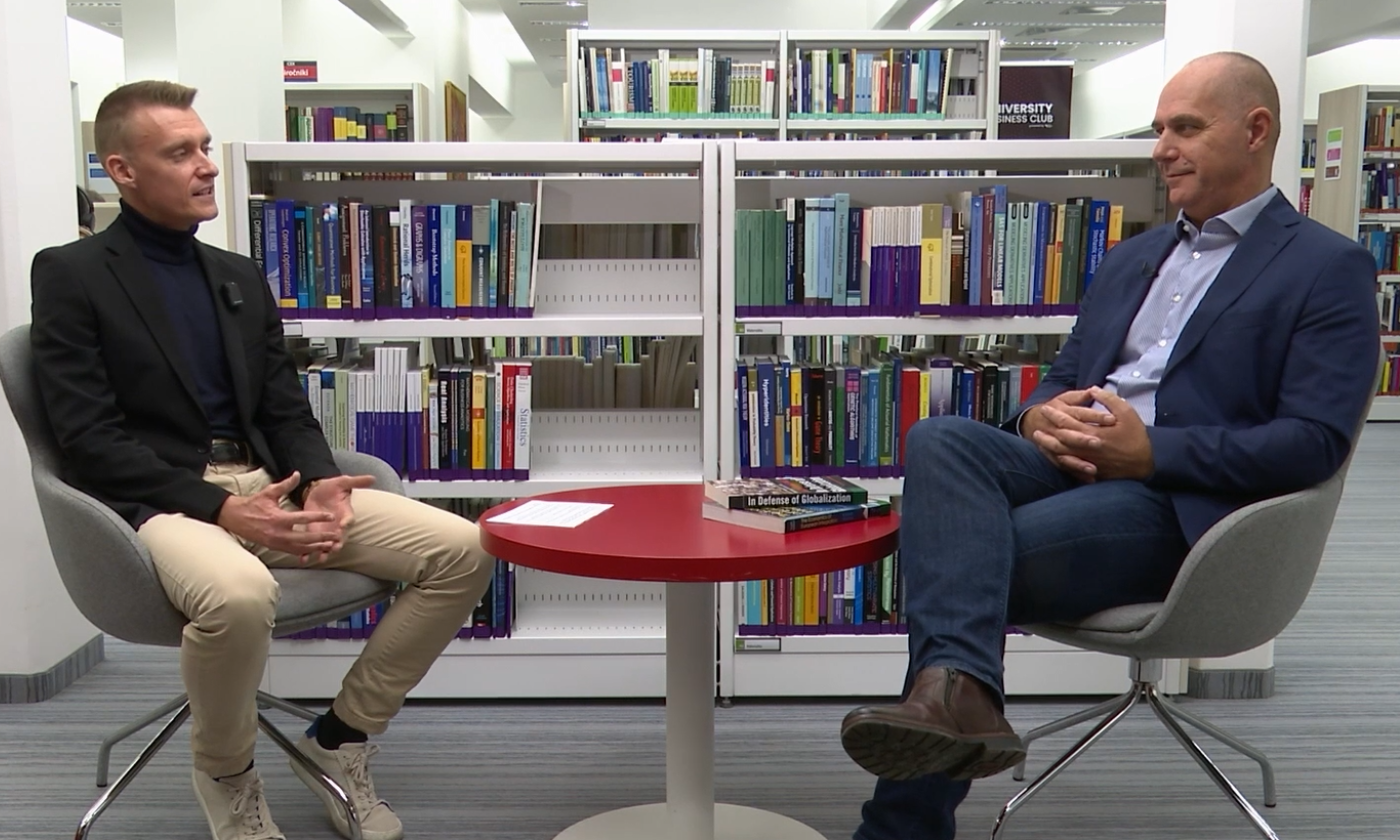 Dva moška sedita na stolih, drug nasproti drugega, v ozadju pa so knjižne police, polne knjig.