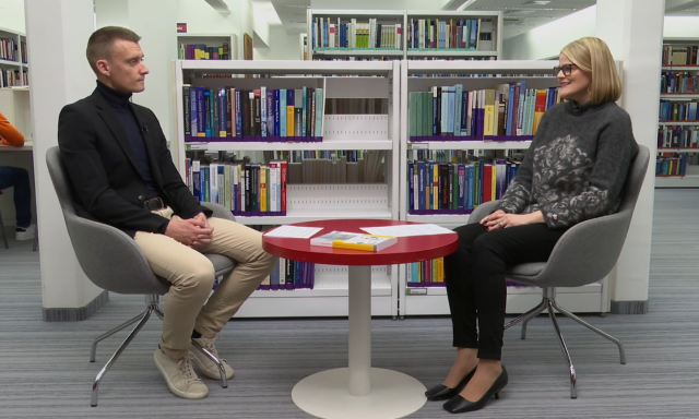 Dve osebi sedita za mizo v knjižnici in se pogovarjata med knjižnimi policami.