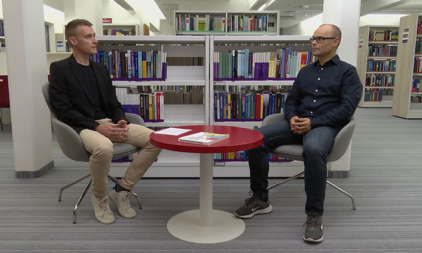 Dva moška sedita drug nasproti drugega v knjižnici, za njima pa so knjižne police. Sedita na sivih stolih okoli majhne rdeče mize, kar nakazuje na poglobljen pogovor.